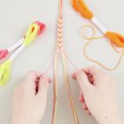 Zwei Hände flechten ein buntes Makramee-Armband. Im Hintergrund sind leuchtende Stickgarnstränge sichtbar.