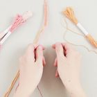 Zu sehen sind zwei Hände, die ein buntes Armband flechten, neben Garnbündeln in Rosa und Orange.