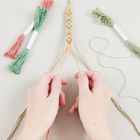 Abgebildet sind Hände, die ein selbst geknüpftes Armband aus farbigen Fäden flechten. Links und rechts liegen Garnbündel.