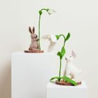 Zwei Hasenfiguren aus Filz, sitzend unter großen, weißen Blumenstängeln mit grünen Blättern, auf weißen Sockeln.