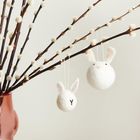 Zwei weiße Filzkugeln mit Hasenohren hängen an Zweigen in einer rosa Vase. Auf einer Kugel ist ein "Y" gestickt.