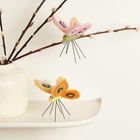 Zwei bunte Filzschmetterlinge mit Drahtbeinen auf weißen Weidenzweigen in einer schlichten Vase.