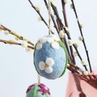 Ein blaues Filzei mit weißen Blumen hängt an Zweigen mit flauschigen Knospen in einer rosafarbenen Vase.