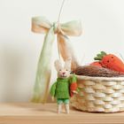 Ein kleines Stoffkaninchen mit grüner Kleidung hält eine Karotte; daneben ein Korb voller lächelnder Karotten.