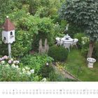 Juli, July, Juillet. Lauschige Sitzgruppe im romantischen Garten. Ein Vogelhaus, blühende Pflanzen, Tisch mit Stühlen im Grünen.