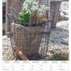 Kalenderblatt April 2025. Drahtkorb mit Holz, Pflanzen. Steine im Hintergrund.