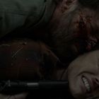 Zwei Personen ringen am Boden: Ein Mann, blutverschmiert, hält die andere Person fest, eine Waffe in der Nähe.