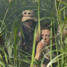 Eine Frau im Vordergrund schleicht aufmerksam durch dichtes, hohes Gras, gefolgt von weiteren Menschen im Hintergrund.