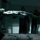 Eine Person in einem weißen Kleid schwebt horizontal in der Luft. Zwei Männer stehen im Hintergrund, Kreuze sind sichtbar.