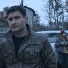 Ein Mann in Lederjacke steht vorne, eine Frau mit Handy neben einem Auto im Hintergrund.