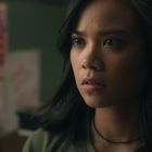 Eine Frau blickt ernst, Hintergrund zeigt Poster und unscharfe Möbel.