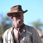 Ein Mann in Westernkleidung, mit braunem Hut und blutigem Gesicht, schaut zur Seite. Im Hintergrund blauer Himmel.