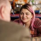 Ich sehe keine erkennbaren Texte. Eine Person mit rosa Haaren lächelt jemanden an, der unscharf im Vordergrund sitzt.