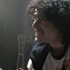 Eine Person mit schwarzer Gesichtsbemalung wird von jemand mit lockigem Haar angesehen, in einem dunklen Raum.