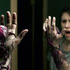 Ein erstaunter Mensch schaut in den Spiegel, die Hand von violetten Adern und einer organischen Kugel bedeckt.