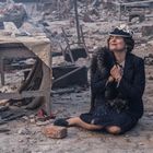 Eine Frau sitzt lächelnd auf Trümmern, hält einen Pelz, umgeben von zerstörten Möbeln und Schutt.