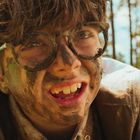 Ein Junge mit Brille und braunen Haaren, Gesicht mit Erde verschmiert, im Freien bei sonnigem Wetter.