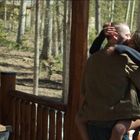 Ein Paar umarmt sich leidenschaftlich auf einer Holzveranda, umgeben von Bäumen. Rechts steht eine Flasche mit Flüssigkeit.