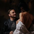 Ein Mann in einer Lederjacke schaut zu einer Frau, die mit dem Rücken zur Kamera steht und ein helles Tuch hält.