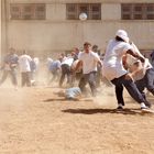 "CDC Prisoner" auf T-Shirts. Menschen kämpfen in einem staubigen Hof mit Sand und Sonne, umgeben von Gebäuden mit Fenstern.