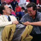 Zwei Männer ringen um einen gelben Blazer auf einem Kleiderbügel und wirken angespannt. Hintergrund: Menschenmenge.