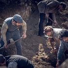 Fünf Männer graben mit Schaufeln und Pickeln in einer sandigen Grube, mit alten, staubigen Kleidern und Mützen.