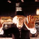 Ein Mann mit wildem, weißem Haar und Hut hebt die Hände, vor einer beleuchteten Stadtkulisse bei Nacht.