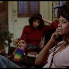 Zwei Frauen in einem Wohnzimmer. Eine telefoniert ernsthaft, die andere sitzt im Hintergrund mit Regenbogenstrümpfen.