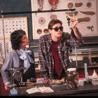 Ein Klassenzimmer mit zwei Personen, ein Mann hält ein Objekt hoch. Text auf einem offenen Buch. Wissenschaftliche Poster.