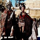 Zwei Personen in aufwendigen, traditionellen Kostümen mit Kopfschmuck und Speer, vor strohgedeckten Strukturen.