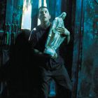 Ich kann keine Texte im Bild erkennen. Ein Mann hält eine religiöse Statue in einem düster beleuchteten Raum.