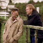 Zwei Männer lehnen am Zaun, einer trägt eine beige Jacke und Mütze, der andere eine schwarze Jacke. Die Landschaft ist ländlich.