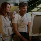 Ein Mann mit farbbekleckstem T-Shirt sitzt vor einer Staffelei; daneben steht eine Frau in einem flauschigen Bademantel.