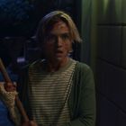 Eine verängstigte Person mit verwuscheltem Haar, blutverschmiert, hält eine lange Stange im diffusen Licht eines Flurs.