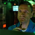 Ein Mann in einem Raum voller Bildschirme mit grünem Licht, in einem blauen Kittel, hält ein Mikrofon.