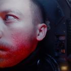 Kein erkennbarer Text. Ein Mann mit rotem Licht im Gesicht sitzt in einem dunklen Raum oder Cockpit, schaut ernst zur Seite.