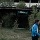 Der Text "REMIS" erscheint auf einem blauen Schild. Im Hintergrund sind Pflanzen und ein altes Gebäude zu sehen.