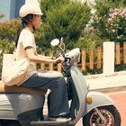 Text: "Verus". Eine Person mit Helm fährt einen grauen Motorroller, umgeben von Bäumen und Blumen.