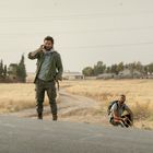 Ein Mann telefoniert stehend auf einer Landstraße, zwei weitere sitzen in einem Feld. Strommasten und Bäume im Hintergrund.