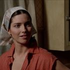 Eine Frau mit einem Kopftuch und rot kariertem Hemd steht in einer Küche mit hölzernen Schränken im Hintergrund.