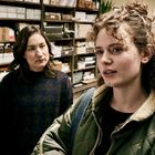 Zwei Frauen stehen in einem Raum mit Regalen voller Kartons und Akten. Eine trägt eine grüne Jacke, die andere einen dunklen Pullover.