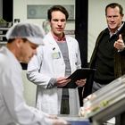 Ein Mann im weißen Kittel hält ein Klemmbrett. Ein anderer Mann zeigt auf etwas und wirkt erklärend. Eine Küche im Hintergrund.