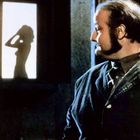 Kein Text sichtbar. Ein Mann betrachtet einen beleuchteten Schattenriss einer Frau im Profil durch ein Fenster.