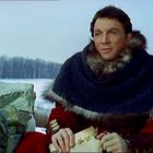 Ein Mann in einem dicken, pelzbesetzten Mantel sitzt draußen bei Schnee. Er hält ein Dokument in der Hand.