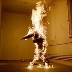 Ein Mensch, vollständig in Flammen, steht in einem kahlen Raum mit gelben Wänden und Metallgitter am Fenster.
