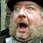 Eine überraschte Person mit offenem Mund und heller Haut, trägt Hut und Mantel, steht vor einer Backsteinmauer.