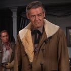 Ein Mann in einem braunen Mantel steht in einem Raum, zwei weitere Personen im Hintergrund. Links ein Porträt, rechts ein Landschaftsbild.