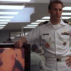 Uniformierter Mann steht neben Fernseher mit abstraktem Bild in einem Büro mit Glaswänden.