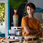 Keine Texte erkennbar. Eine Frau in einem gelben Shirt steht mit verschränkten Armen in einer Küche.