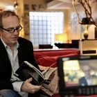Ein Mann mit Brille sitzt auf einem roten Sofa, liest ein Buch, im Hintergrund unscharfe Wohnzimmerdetails.